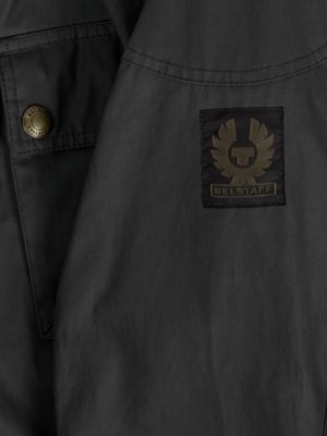 Nahaufnahme einer schwarzen Jacke. Auf der Jacke befinden sich ein runder Knopf, eine Brusttasche und ein Aufnäher mit dem Logo von Belstaff in Gold auf Schwarz. Der Stoff ist glatt und leicht glänzend.
