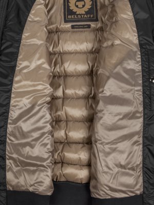 Nahaufnahme des Inneren einer schwarzen, gesteppten Jacke. Das Innenfutter ist hellbraun. Oben ist ein Label mit "BELSTAFF" und Logo zu sehen. Die Jacke ist offen.
