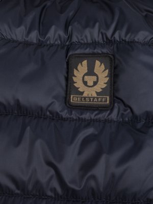 Detailaufnahme einer dunkelblauen, gesteppten Jacke mit einem aufgenähten Belstaff-Emblem. Das Emblem zeigt einen goldenen, geflügelten Schriftzug "Belstaff" in einem rechteckigen Feld. Die Steppnähte verlaufen horizontal.