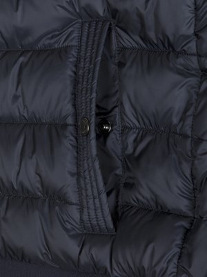 Nahaufnahme einer dunkelblauen, gesteppten Jacke mit einer aufgesetzten Tasche. Die Tasche hat einen Knopfverschluss. Der Stoff der Jacke ist glänzend. Die Naht der Tasche ist vertikal verlaufend.