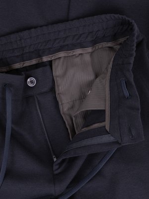 Nahaufnahme einer geöffneten dunklen Hose mit Reißverschluss und Knopf. Der Bund der Hose ist gerippt. Die Innenseite der Hose ist grau. Ein Kordelzug ist sichtbar. Die Hose ist aus Stoff gefertigt.
