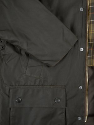 Naheinstellung einer dunkelgrünen Jacke. Ein Ärmel, der Teil der Jacke ist, ragt nach links. Ein rechteckige Tasche befindet sich im unteren Bereich mit zwei Knöpfen. Ein Reißverschluss ist sichtbar. Ein kariertes Innenfutter ist zu sehen.