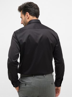 Ein Mann mit dunklem Haar, von hinten fotografiert, trägt ein schwarzes Hemd mit Kragen und Manschettenärmeln. Er hat eine graue Hose an. Sein Kopf ist leicht zur Seite geneigt. Der Hintergrund ist weiß.