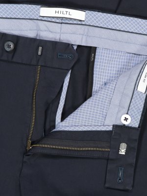 Nahaufnahme einer geöffneten dunklen Hose. Der Reißverschluss ist geöffnet, die Knopfleiste und der Bund sind sichtbar. Das Innenfutter ist hellblau mit einem geometrischen Muster. Ein Etikett mit der Aufschrift "HILTL" ist zu sehen.