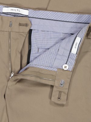 Nahaufnahme einer geöffneten beigefarbenen Hose mit Reißverschluss und Knopf. Innenfutter mit kariertem Muster. Auf einem Etikett steht "SLIM FIT". Über dem Bund ist "HILTL" aufgedruckt. Das Innenfutter ist hellblau.