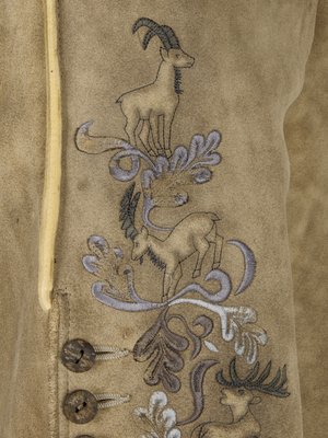 Naheinstellung einer hellbraunen Lederhose. Sie ist mit gestickten Steinböcken und Hirsch verziert. Zwei Knöpfe sind sichtbar. Links ist ein gelber Streifen als Kontrast zu sehen.