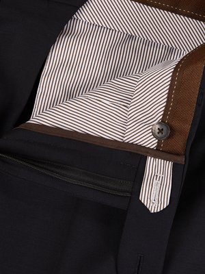Nahaufnahme einer schwarzen Hose. Sichtbar sind der Hosenbund, ein Knopf und der Reißverschluss. Das Innenfutter weist ein Muster aus braunen und weißen Streifen auf. Der Saum ist mit braunem Stoff eingefasst.