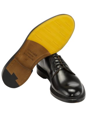 Nahaufnahme eines schwarzen Lederschuhs mit Schnürsenkeln. Der Schuh ist leicht von unten gezeigt, wobei die gelbe Sohle und die braune Innensohle sichtbar sind. Auf der gelben Sohle steht Doucal's.