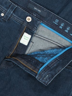 Nahaufnahme einer Jeans in dunklem Denim-Blau. Der Reißverschluss ist offen und die Innenseite mit blauem Stoff und Muster ist sichtbar. Oben ein Knopf, ein Label mit "pierre cardin" und Icons. Ein grünes "Green Rivet"-Etikett ist auch zu sehen.