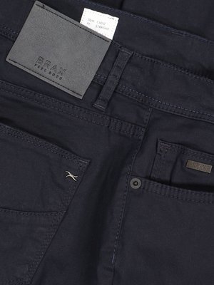 Nahaufnahme einer dunkelblauen Jeanshose. Oben links ein graues Lederetikett mit "BRAX FEEL GOOD".  Darunter befindet sich ein weißes Etikett mit Produktinformationen. Auf der rechten Seite sind Gesäßtaschen und ein Knopf sichtbar.