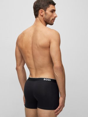 Ein Mann, von hinten und der Seite gesehen, steht vor hellem Hintergrund. Er trägt schwarze Boxershorts mit weißer Schrift "BOSS" am Bund. Sein Oberkörper ist unbekleidet und er schaut zur Seite.