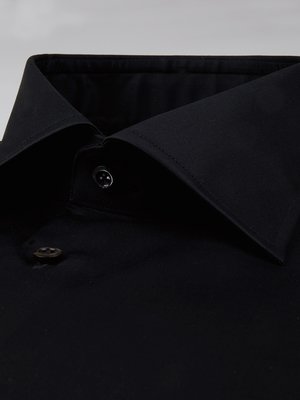 Nahaufnahme des Kragens und der Knopfleiste eines schwarzen Hemdes. Der Kragen ist leicht abgewinkelt, und zwei Knöpfe sind sichtbar. Der Stoff wirkt glatt und leicht glänzend. Der Hintergrund ist hell und unscharf.