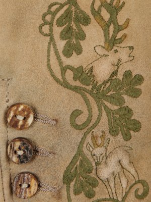Beige Lederimitat mit grüner Stickerei in Form von Blättern und zwei Rehen. Auf der linken Seite sind drei runde Knöpfe aus Naturmaterialien befestigt. Rechts ist die Stickerei von einem Hirsch mit Geweih zu sehen.