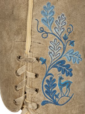 Nahaufnahme einer beigefarbenen Lederjacke mit blauer Stickerei. Auf der Jacke ist ein blaues Blattmuster mit einem Reh gestickt. Auf der linken Seite befindet sich eine Schnürung mit hellen Schnüren.