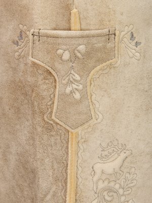 Nahaufnahme einer beigefarbenen Wildlederhose mit dunkelgrauen Stickereien. Dargestellt sind eine Tasche mit Eicheln und Blättern sowie ein Hirsch und Blätter. Die Hosennähte sind sichtbar.