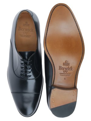 Zwei schwarze Lederschuhe, einer von oben, einer von unten betrachtet. Der linke Schuh ist geschnürt, der rechte zeigt die Sohle mit dem Markenlogo "Berwick 1707". Die Sohle hat eine braune Farbe und ist mit einer Naht versehen.