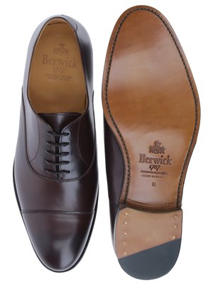 Zwei braune Lederschuhe, einer von oben und einer von unten betrachtet. Der rechte Schuh zeigt die Sohle, auf der "Berwick 1707" und "GOOD YEAR WELTED" steht. Beide Schuhe sind Schnürschuhe.