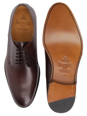 Zwei braune Lederschuhe, links ein Schnürschuh mit Schnürsenkeln, rechts die Schuhsohle mit Prägung. Die Schuhsohle ist hellbraun, mit schwarzem Gummiauftrag an der Spitze.