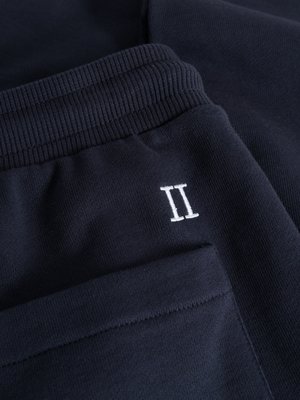 Nahaufnahme einer dunkelblauen Hose aus Sweatstoff. Der Fokus liegt auf der linken Hosentasche und dem Hosenbund. Auf der Hose ist ein weißes "II" gestickt. Der Stoff wirkt weich und hat eine gerippte Struktur am Bund.