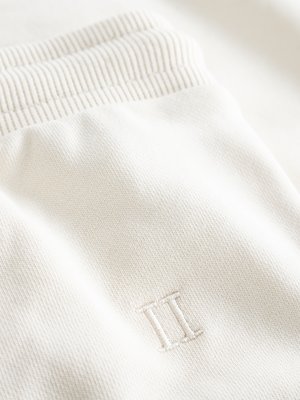 Nahaufnahme eines Kleidungsstücks in Off-White. Oben ist ein gerippter Bund zu sehen, darunter der Stoff mit einem eingestickten Logo mit zwei parallelen vertikalen Strichen. Der Stoff weist eine feine Textur auf.