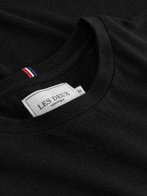 Nahaufnahme eines schwarzen T-Shirts mit Rundhalsausschnitt. Auf dem Kragen ist ein kleines rot-weiß-blaues Detail. Ein Etikett mit "LES DEUX copenhagen" ist angenäht. Daneben steht ein "M".