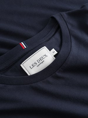 Nahaufnahme eines dunkelblauen T-Shirts. Das Shirt hat einen Rundhalsausschnitt und ein kleines Etikett mit der Aufschrift "LES DEUX Copenhagen". Auf dem Nackenband befinden sich rote, weiße und blaue Streifen. Das Etikett hat eine Größe "M".