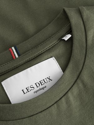 Nahaufnahme eines olivgrünen T-Shirts. Der Rundhalsausschnitt ist erkennbar. Ein weißes Etikett mit dem Markennamen "LES DEUX copenhagen" ist sichtbar. Am Nacken befinden sich drei kleine Streifen: rot, weiß und blau.