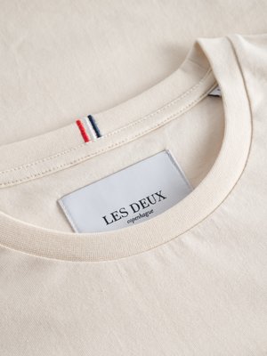 Nahaufnahme eines beigen T-Shirts mit Rundhalsausschnitt. Auf dem Kragen sind drei farbige Streifen, rot, blau und weiß, gestickt. Aufgenäht ist ein weißes Label mit der Aufschrift "LES DEUX copenhagen".