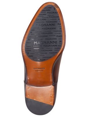 Nahaufnahme eines braunen Lederschuhs. Die Sohle zeigt ein geprägtes Muster mit dem Schriftzug "MAGNANNI" auf grauem Hintergrund. Der Schuhabsatz ist ebenfalls braun und hat eine helle Verstärkung mit metallischen Elementen.