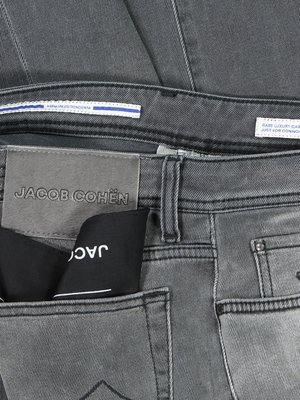 Nahaufnahme einer grauen Jeans mit dem Label "JACOB COHEN" an der Gesäßtasche. Eine schwarze Tasche mit weißem "JAC" ragt aus der Tasche. Ein weißes Etikett mit blauer Linie und Text ist oben zu sehen. Die Jeans ist gefaltet.