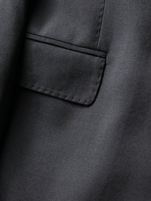 Nahaufnahme eines schwarzen Anzugs. Der Fokus liegt auf der Brusttasche, die durch eine abgesteppte Naht vom Rest des Stoffes abgegrenzt ist. Der Stoff weist eine feine Textur auf. Die Oberflächen des Anzugs sind dunkelgrau und glatt.