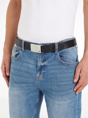 Ein Mann trägt eine Jeans und ein weißes T-Shirt. Er hält sich mit seinen Händen fest. Die Jeans hat eine blaue Farbe und einen schwarzen Gürtel mit einer silbernen Schnalle. Der Gürtel ist auf seiner Taille befestigt.