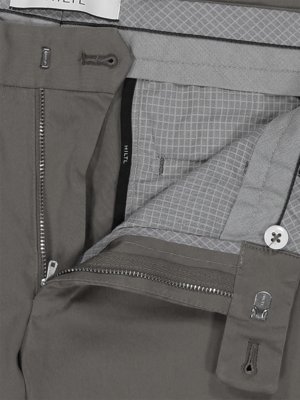 Nahaufnahme einer geöffneten grauen Hose. Die Hose zeigt eine offene Reißverschlussleiste, einen Knopf und eine Innentasche aus grauem Stoff mit einem karierten Muster. Ein Etikett mit dem Wort "HILTL" ist sichtbar.