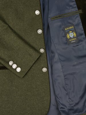 Nahaufnahme eines dunkelgrünen Sakkos. Der Kragen hat eine dunkle Paspelierung. Das Innenfutter ist blau, ein Etikett mit dem Wappen Bayerns ist sichtbar. Silberfarbene Knöpfe an der Knopfleiste und Ärmel.