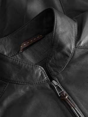 Nahaufnahme des Kragens und Reißverschlusses einer schwarzen Lederjacke. Der Kragen ist rund und die Jacke ist mit Ziernähten versehen. Der Reißverschluss ist geöffnet und hat einen braunen Lederanhänger.