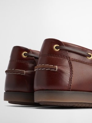 Nahaufnahme von zwei braunen Lederschuhen. Die Rückseite der Schuhe ist zu sehen, mit Ösen aus Metall und braunen Schnürsenkeln. Auf dem Fersenbereich sind helle Nähte. Die Sohlen sind braun und leicht abgerundet.
