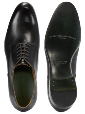 Ein Paar schwarze Lederschuhe, eines mit der Oberseite und eines mit der Sohle nach oben. Der Schuh auf der linken Seite ist geschnürt. Auf der Sohle steht SCHUHE & HANDWERK.