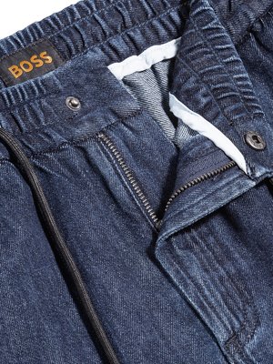 Nahaufnahme einer Jeanshose in Blau. Die Hose hat einen Reißverschluss, einen Knopf und eine Kordel. Ein BOSS-Logo-Etikett ist auf dem Bund zu sehen. Der Stoff ist strukturiert. Die Hose ist offen.