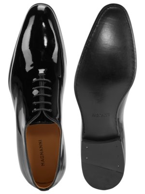 Zwei schwarze Lederschuhe auf weißem Hintergrund. Der linke Schuh ist ein Schnürschuh mit glänzendem Leder. Der rechte Schuh ist umgedreht, die Sohle ist sichtbar. Auf der Sohle steht "MAGNANNI".