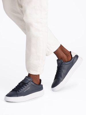 Nahaufnahme eines Beines in weißen Hosen und blauen Sneakern mit weißer Sohle. Die Schuhe haben Schnürsenkel und das Tommy Hilfiger Logo. Der Hintergrund ist weiß.
