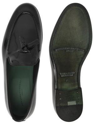 Zwei schwarze Lederslipper, einer mit sichtbarer Oberseite, der andere mit der Unterseite nach oben. Der linke Slipper hat eine Quaste und eine grüne Innensohle. Der rechte hat eine grüne Sohle mit Etiketten.