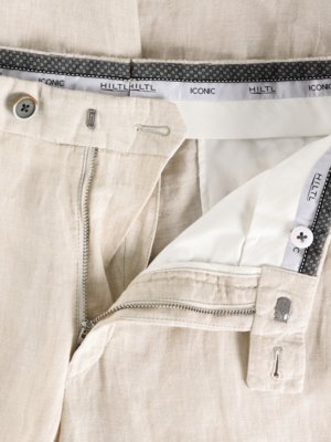 Nahaufnahme einer geöffneten beigen Hose aus Leinen. Der Reißverschluss ist geöffnet, Knöpfe und Knopflöcher sind sichtbar. Ein Stoffband mit Schriftzug „HILTL“ und „ICONIC“ umrandet die Hose.