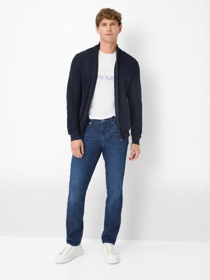 Ein Mann steht vor weißem Hintergrund und trägt einen marineblauen Strick-Cardigan, ein weißes T-Shirt und blaue Jeans. Er trägt weiße Sneakers. Sein Blick geht in die Kamera.