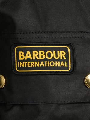 Nahaufnahme einer schwarzen Stoffoberfläche mit einem rechteckigen Aufnäher. Auf dem Aufnäher steht in gelber Schrift "BARBOUR INTERNATIONAL" auf schwarzem Grund. Links und rechts sind goldene Knöpfe zu sehen.