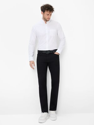 Ein Mann steht vor weißem Hintergrund. Er trägt ein weißes Hemd mit Knöpfen, schwarze Jeans, einen schwarzen Gürtel und weiße Sneakers. Er hat lockiges Haar und blickt nach unten. Sein linker Arm ist in der Hosentasche.