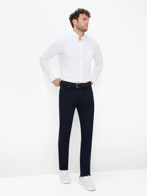 Ein Mann steht vor weißem Hintergrund und trägt ein weißes Hemd, eine blaue Jeans und weiße Turnschuhe. Er hat lockiges, braunes Haar und einen schwarzen Gürtel. Seine Hände sind in seine Hosentaschen gesteckt.