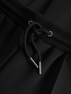 Nahaufnahme einer schwarzen Hose mit einem schwarzen Kordelzug, der durch Metallösen geführt ist. Der Kordelzug ist in der Mitte geknotet. Die Enden des Kordelzugs sind mit silberfarbenen Kappen versehen. Der Stoff ist glatt und leicht glänzend.