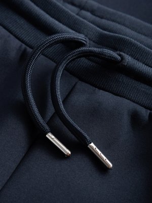 Nahaufnahme einer dunkelblauen Hose. Oben sieht man ein gekräuseltes, dunkles Seil, das durch zwei metallische Ösen geführt wird. Der Hintergrund zeigt Stoff mit feinen vertikalen Linien.