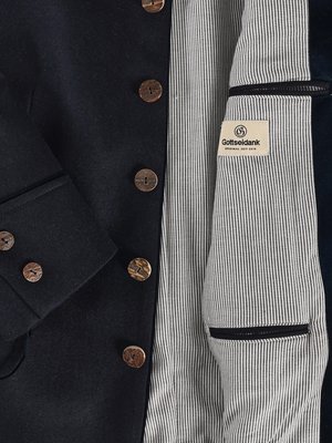 Nahaufnahme einer dunklen Jacke mit Knöpfen. Innenfutter ist weiß mit feinen, vertikalen, schwarzen Streifen. Auf einem Etikett steht "Gottseidank". Die Jacke hat Holzknöpfe und eine Innentasche.