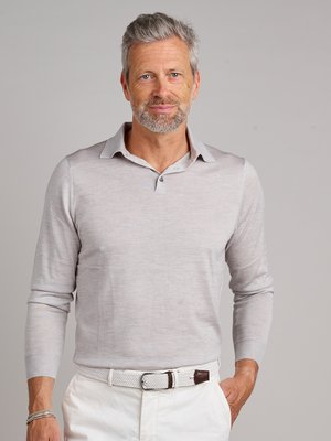 Ein Mann mittleren Alters mit grauem Haar und Bart trägt ein hellgraues Langarm-Polo-Shirt. Er hat eine silberne Armband am linken Handgelenk, und eine weiße Hose mit weißem geflochtenem Gürtel mit braunen Details. Der Hintergrund ist hellgrau.
